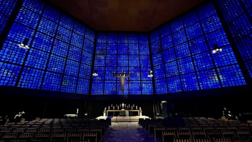 A large, modern church interior with walls composed of square blue stained glass panels. A crucifix is centrally positioned above the altar. Rows of empty chairs are arranged neatly, facing the altar.