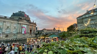 A lively outdoor gathering near a historic building with a dome. There are numerous people socializing and enjoying the event under strings of lights at sunset. The sky is vibrant with hues of orange, pink, and blue, providing a picturesque backdrop.