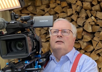 A man with glasses is operating a professional video camera in front of a stack of chopped wood logs. He is wearing a light blue shirt and red suspenders, with gray hair, and is looking slightly upward.