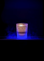 A lit candle with a label that reads 'Only For You, Handmade Candle, Aromatherapy Experts' is placed against a dark background. The candle emits a soft, warm glow surrounded by a subtle blue mist or smoke.