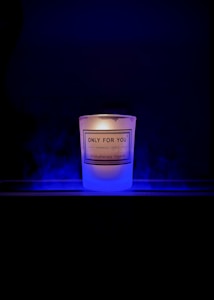 A lit candle with a label that reads 'Only For You, Handmade Candle, Aromatherapy Experts' is placed against a dark background. The candle emits a soft, warm glow surrounded by a subtle blue mist or smoke.