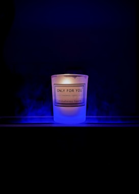 A lit candle with a label that reads 'Only For You, Handmade Candle, Aromatherapy Experts' is placed against a dark background. The candle emits a soft, warm glow surrounded by a subtle blue mist or smoke.