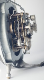 An intricate close-up of a mechanical clock or watch mechanism, showcasing interlocking gears and metal components with a focus on the detailed craftsmanship. The image features a soft focus and muted lighting, highlighting the metal textures and aged appearance.