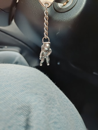 A small astronaut figurine keychain is hanging from a metal chain attached to a key inserted in an ignition. The interior of a car is visible, including part of the dashboard and seat. A fabric with a light blue tint is in the foreground.