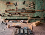 A woodworking workshop with various tools neatly arranged on wall-mounted shelves. The tools include saws, files, clamps, and hand planes. The workbench is cluttered with wooden planks, chisels, a mallet, and shavings. The background features a textured green wall, providing a rustic atmosphere.