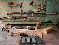 A woodworking workshop with various tools neatly arranged on wall-mounted shelves. The tools include saws, files, clamps, and hand planes. The workbench is cluttered with wooden planks, chisels, a mallet, and shavings. The background features a textured green wall, providing a rustic atmosphere.