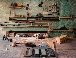 A woodworking workshop with various tools neatly arranged on wall-mounted shelves. The tools include saws, files, clamps, and hand planes. The workbench is cluttered with wooden planks, chisels, a mallet, and shavings. The background features a textured green wall, providing a rustic atmosphere.