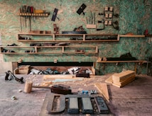 A woodworking workshop with various tools neatly arranged on wall-mounted shelves. The tools include saws, files, clamps, and hand planes. The workbench is cluttered with wooden planks, chisels, a mallet, and shavings. The background features a textured green wall, providing a rustic atmosphere.