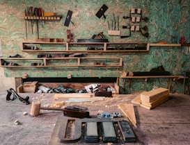 A woodworking workshop with various tools neatly arranged on wall-mounted shelves. The tools include saws, files, clamps, and hand planes. The workbench is cluttered with wooden planks, chisels, a mallet, and shavings. The background features a textured green wall, providing a rustic atmosphere.