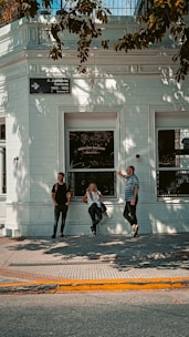 Three people stand in front of a white building with the name 'Mercado McCann' visible on one of the windows. The building has intricate architectural details and a small street sign. Shadows of tree branches cast patterns on the building facade.