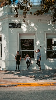 Three people stand in front of a white building with the name 'Mercado McCann' visible on one of the windows. The building has intricate architectural details and a small street sign. Shadows of tree branches cast patterns on the building facade.