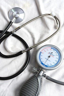 A stethoscope and a sphygmomanometer are laid out on a white fabric surface. The stethoscope has a shiny metallic diaphragm and black tubing. The sphygmomanometer features a round gauge with blue, red, and black markings indicating blood pressure measurements, attached to a gray and black bulb used for inflating the cuff.