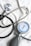 A stethoscope and a sphygmomanometer are laid out on a white fabric surface. The stethoscope has a shiny metallic diaphragm and black tubing. The sphygmomanometer features a round gauge with blue, red, and black markings indicating blood pressure measurements, attached to a gray and black bulb used for inflating the cuff.