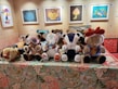 A collection of plush teddy bears with unique clothing and designs are displayed on a decorative tablecloth with floral patterns. Behind them, several framed artworks hang on the wall including images of a cartoon character and various colorful abstract designs. One bear wears goggles and another is adorned with stickers. A plush camel toy lies beside the bears.