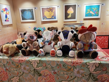 A collection of plush teddy bears with unique clothing and designs are displayed on a decorative tablecloth with floral patterns. Behind them, several framed artworks hang on the wall including images of a cartoon character and various colorful abstract designs. One bear wears goggles and another is adorned with stickers. A plush camel toy lies beside the bears.