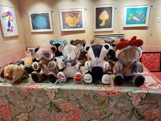 A collection of plush teddy bears with unique clothing and designs are displayed on a decorative tablecloth with floral patterns. Behind them, several framed artworks hang on the wall including images of a cartoon character and various colorful abstract designs. One bear wears goggles and another is adorned with stickers. A plush camel toy lies beside the bears.