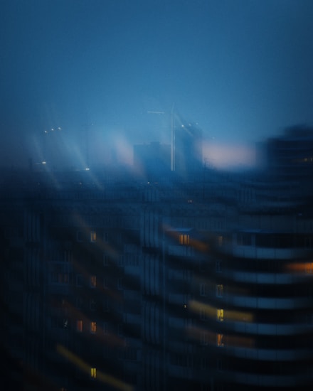 A cityscape with a blurred, dreamy appearance during nighttime or dusk. The image features large, shadowy buildings with small lit windows, and a foggy, atmospheric skyline in the background. Streetlights and faint structural outlines add to the ethereal mood.