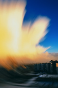 A blurred image featuring a dramatic sky displaying a large, luminous cloud formation transitioning from yellow to white against a backdrop of deep blue. Below the sky, there are a cluster of buildings, suggesting a cityscape or residential area.