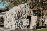 A small structure resembling a house built entirely out of open books. The books are stacked over frame-like walls, creating an artistic installation. The setting appears to be an outdoor area, likely a plaza, with trees and building facades visible in the background. There is a door on the structure and posters or signs attached to it.