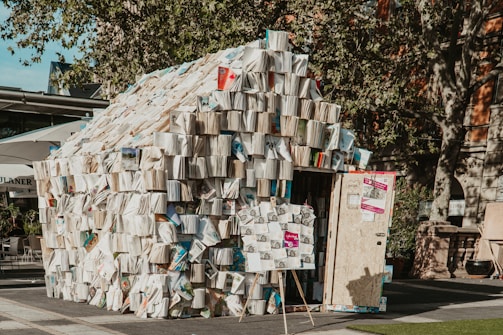 A small structure resembling a house built entirely out of open books. The books are stacked over frame-like walls, creating an artistic installation. The setting appears to be an outdoor area, likely a plaza, with trees and building facades visible in the background. There is a door on the structure and posters or signs attached to it.