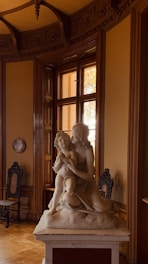 A sculpture depicting two figures, one appearing to embrace the other, is prominently displayed on a pedestal in a richly decorated room. The room features ornate woodwork and design elements, with high ceilings and a window letting in natural light. Two antique chairs are positioned near the walls, completing the classic and elegant ambiance.