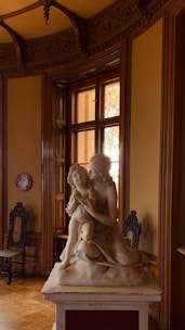 A sculpture depicting two figures, one appearing to embrace the other, is prominently displayed on a pedestal in a richly decorated room. The room features ornate woodwork and design elements, with high ceilings and a window letting in natural light. Two antique chairs are positioned near the walls, completing the classic and elegant ambiance.