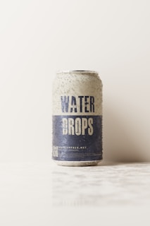 A soda can with the words 'Water Drops' is standing upright on a white surface. The can has a textured appearance with water droplets on its exterior. The upper part of the can is light, while the bottom half is dark blue. The background is plain and light-colored, giving a minimalistic look.