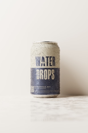 A soda can with the words 'Water Drops' is standing upright on a white surface. The can has a textured appearance with water droplets on its exterior. The upper part of the can is light, while the bottom half is dark blue. The background is plain and light-colored, giving a minimalistic look.
