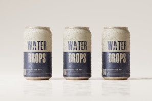 Three beverage cans covered in condensation are aligned in a row against a light, neutral background. The cans are labeled with the words 'Water Drops' in bold blue letters on a white background, with additional text and a QR code near the bottom.