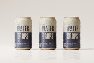 Three beverage cans covered in condensation are aligned in a row against a light, neutral background. The cans are labeled with the words 'Water Drops' in bold blue letters on a white background, with additional text and a QR code near the bottom.