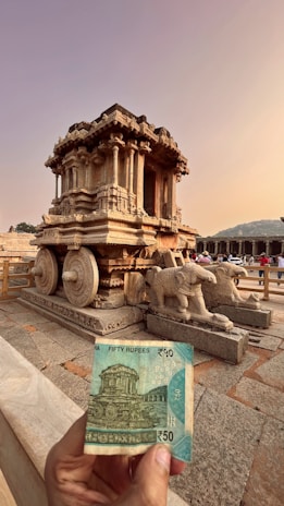 A stone chariot structure in an ancient architectural style with intricate carvings is prominently displayed. In the foreground, a hand holds a fifty rupee Indian banknote featuring a depiction of the same chariot, highlighting the resemblance between the two. The setting is outdoor with a clear sky and distant views of a mountain range and some surrounding structures.
