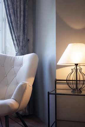 A cozy living room corner with beige and muted brown textiles layered on a comfy armchair beside a softly glowing lamp.