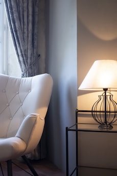 A cozy room corner featuring a tufted white armchair near a softly lit lamp. The lamp rests on a black metal side table with a glass top. A window with floral-patterned drapes is visible in the background, letting in natural light.