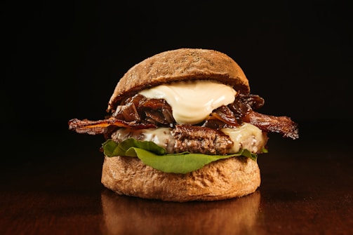 A gourmet burger is presented with a dark, glossy bun and layers of crispy bacon, melted cheese, and caramelized onions. The juicy beef patty sits atop fresh green lettuce, topped with a dollop of creamy sauce. The warm lighting creates a rich, inviting atmosphere, highlighting the textures and colors of the ingredients.