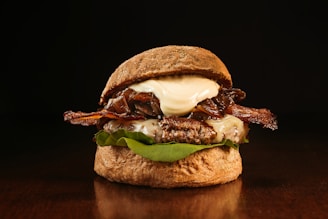 A gourmet burger is presented with a dark, glossy bun and layers of crispy bacon, melted cheese, and caramelized onions. The juicy beef patty sits atop fresh green lettuce, topped with a dollop of creamy sauce. The warm lighting creates a rich, inviting atmosphere, highlighting the textures and colors of the ingredients.