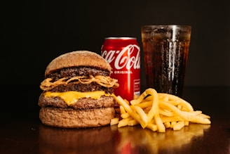 A large burger with layered beef patties, cheese, and fried onions sits on a dark surface. Next to it are golden crispy french fries. In the background, a chilled can of Coca-Cola and a tall glass filled with a cola beverage, topped with ice, create a classic fast food meal setting.