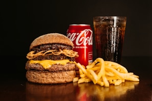 A large burger with layered beef patties, cheese, and fried onions sits on a dark surface. Next to it are golden crispy french fries. In the background, a chilled can of Coca-Cola and a tall glass filled with a cola beverage, topped with ice, create a classic fast food meal setting.