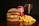 A large burger with layered beef patties, cheese, and fried onions sits on a dark surface. Next to it are golden crispy french fries. In the background, a chilled can of Coca-Cola and a tall glass filled with a cola beverage, topped with ice, create a classic fast food meal setting.