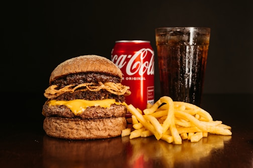 A large burger with layered beef patties, cheese, and fried onions sits on a dark surface. Next to it are golden crispy french fries. In the background, a chilled can of Coca-Cola and a tall glass filled with a cola beverage, topped with ice, create a classic fast food meal setting.