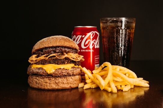 A large burger with layered beef patties, cheese, and fried onions sits on a dark surface. Next to it are golden crispy french fries. In the background, a chilled can of Coca-Cola and a tall glass filled with a cola beverage, topped with ice, create a classic fast food meal setting.