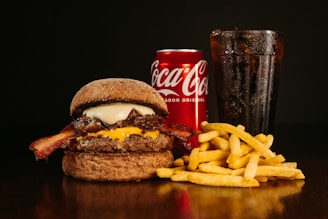 A appetizing hamburger with melted cheese, crispy bacon, and savory sauce sits beside a generous serving of golden fries. In the background, a red Coca-Cola can and a glass of the beverage filled with ice complement the meal.