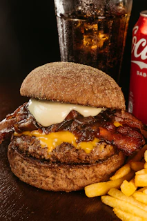 Close-up of a juicy burger with crispy fries and a cold soda on a red and black table.