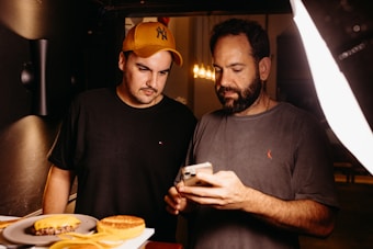 Two men are focused on a smartphone, possibly discussing or reviewing something on the screen. There are plates of food, including a burger and a dessert, on the table nearby. One man is wearing a yellow cap and a black shirt, while the other is bearded and wearing a grey shirt.