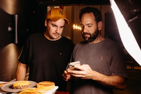 Two men are focused on a smartphone, possibly discussing or reviewing something on the screen. There are plates of food, including a burger and a dessert, on the table nearby. One man is wearing a yellow cap and a black shirt, while the other is bearded and wearing a grey shirt.