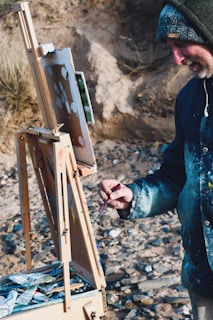 A person is painting outdoors, using an easel set on a rocky beach. The individual is wearing a hooded jacket and holding a paintbrush. The painting appears to be in progress with various colors layered on the canvas. Around the easel, there are tubes of paint and art supplies scattered in a wooden box.