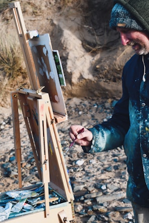 A person is painting outdoors, using an easel set on a rocky beach. The individual is wearing a hooded jacket and holding a paintbrush. The painting appears to be in progress with various colors layered on the canvas. Around the easel, there are tubes of paint and art supplies scattered in a wooden box.