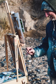 A person is painting outdoors, using an easel set on a rocky beach. The individual is wearing a hooded jacket and holding a paintbrush. The painting appears to be in progress with various colors layered on the canvas. Around the easel, there are tubes of paint and art supplies scattered in a wooden box.