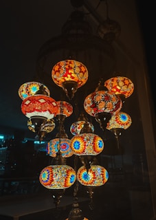A collection of intricately designed hanging lamps with vibrant mosaic patterns, featuring colors like blue, red, orange, and yellow. The lamps create a warm and inviting atmosphere against a dark background.