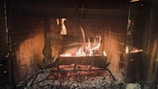 A warm and cozy fire burns brightly within a fireplace, surrounded by a protective mesh screen. The flames cast a gentle glow, illuminating the brick interior of the fireplace. Logs are arranged on a metal grate, contributing to the flickering firelight and emitting a comforting ambiance.