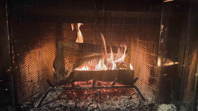 A warm and cozy fire burns brightly within a fireplace, surrounded by a protective mesh screen. The flames cast a gentle glow, illuminating the brick interior of the fireplace. Logs are arranged on a metal grate, contributing to the flickering firelight and emitting a comforting ambiance.